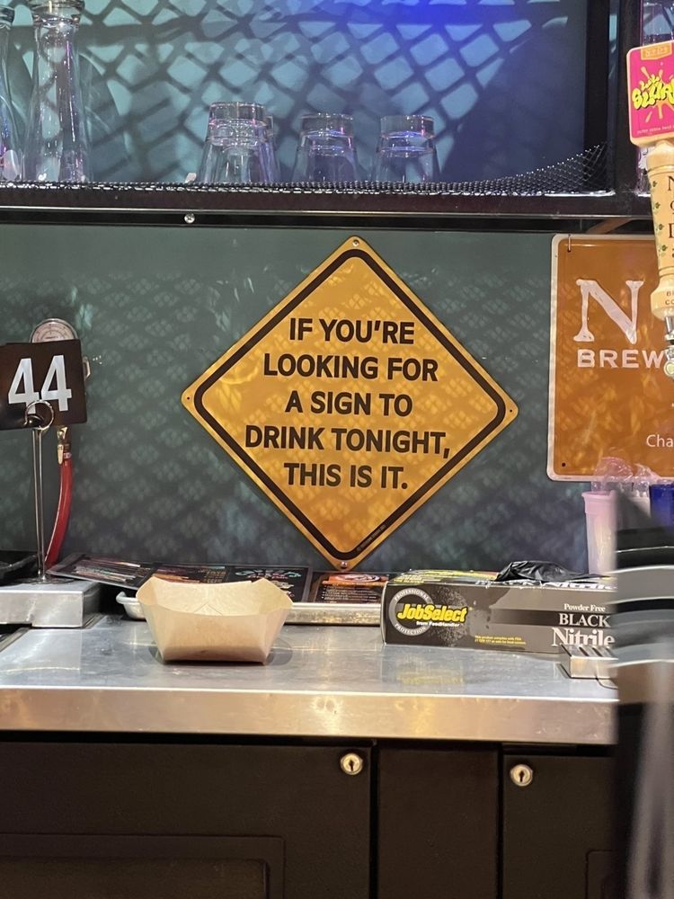 A fake traffic warning sign over a bar next to beer taps that tears “If you’re looking for a sign to drink tonight, this is it”