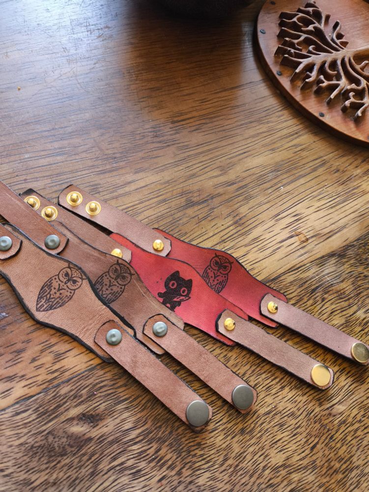 Quatro pulseiras, duas marrons e duas meio vermelhas. Em três há uma coruja desenhada. E uma delas um gatinho com uma faca na mão. 