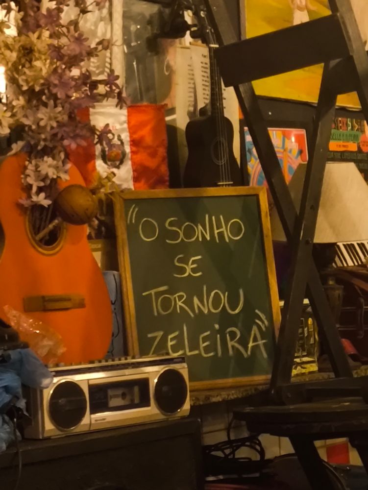 Imagem de um quadro verde escrito O sonho se tornou zeleira. Ao lado tem um violão e um rádio. Do outro lado tem uma cadeira. 