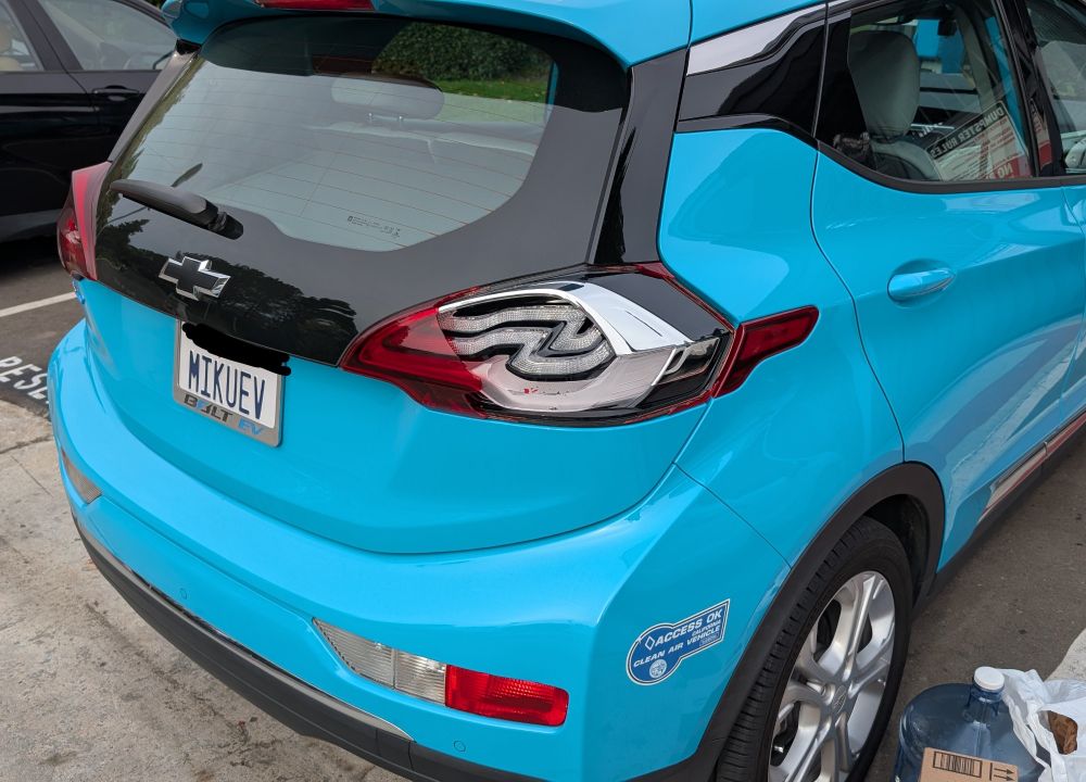 A turquoise Chevrolet Bolt EV with one of its red tail lights smashed. The car has the same color scheme as Hatsune Miku.