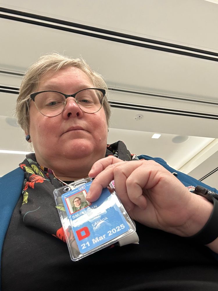 Vicky holds a blue UN badge. She is a middle aged woman with short blonde and gray hair and glasses, wearing a teal jacket and a floral scarf 
