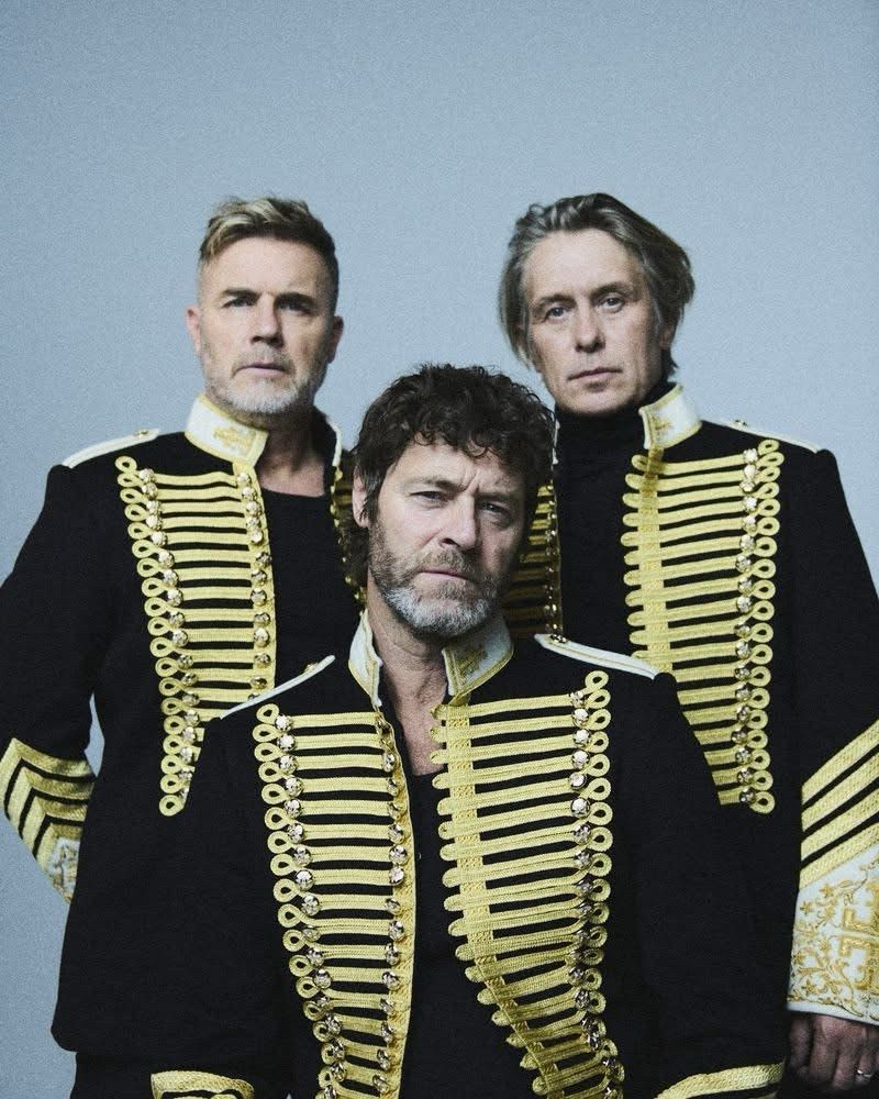 Gary Barlow (l), Howard Donald (middle) and Mark Owen (r) dressed in Adam Ant coats looking moody; Barlow and Donald have clipped beards.