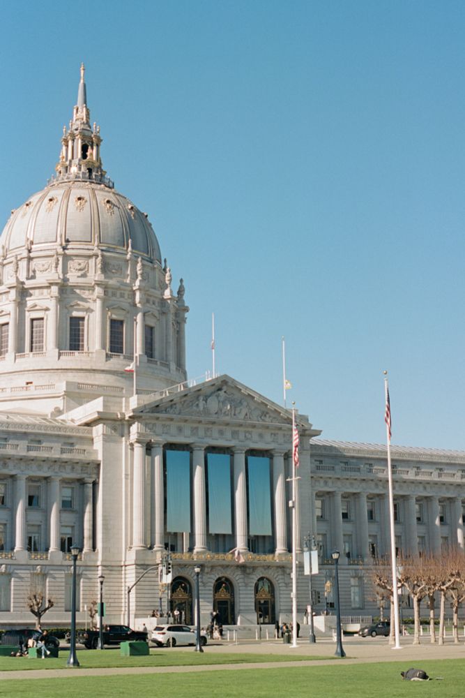 A picture of City Hall in San Francisco, California. A person sleeps in the grass in front of the building. 