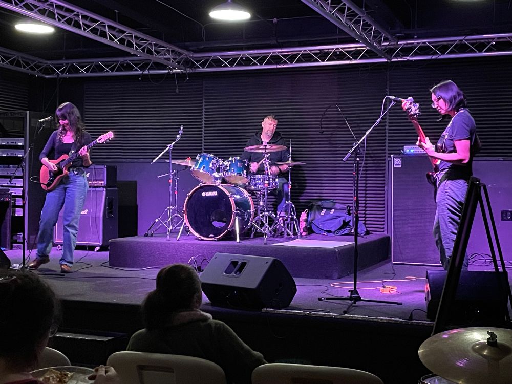 A rock band on a stage comprising a guitarist, drummer, and bassist.