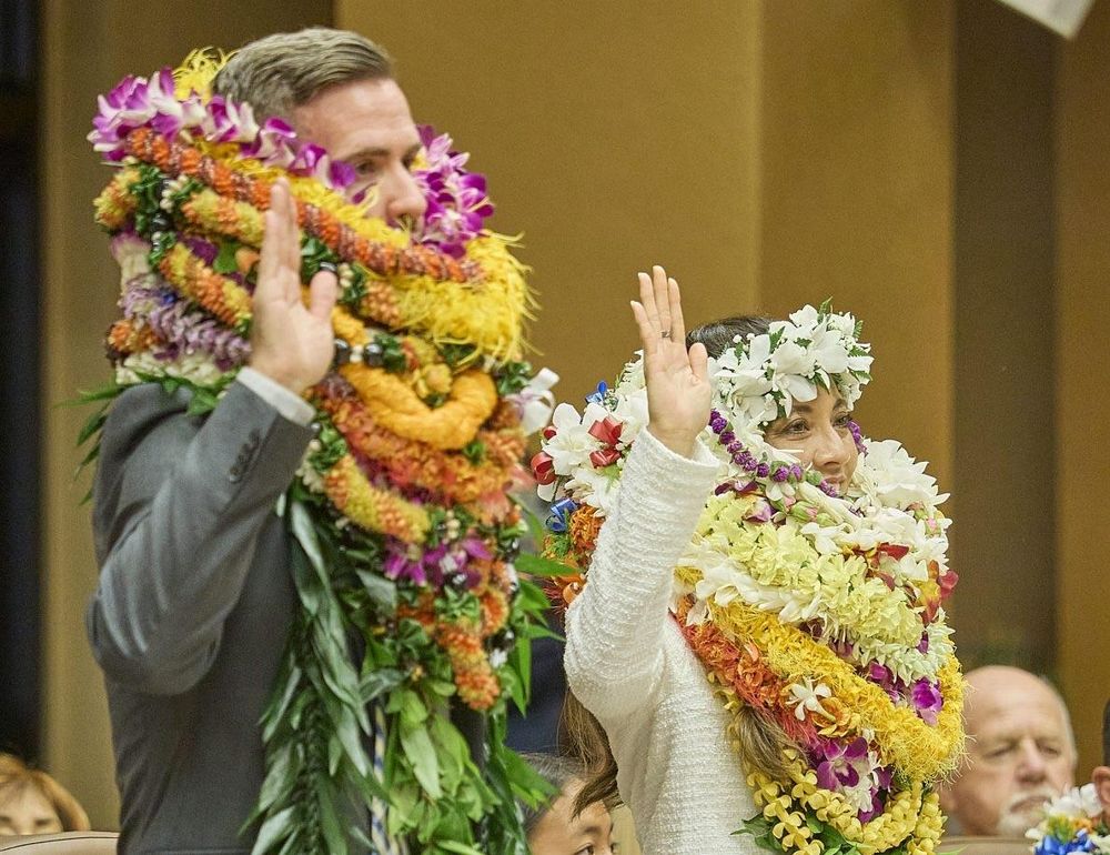 A man and woman are raising their right hands in a ceremonial setting, adorned with vibrant floral lei. The man is dressed in a suit, while the woman wears a white outfit. In the background, spectators watch the event.