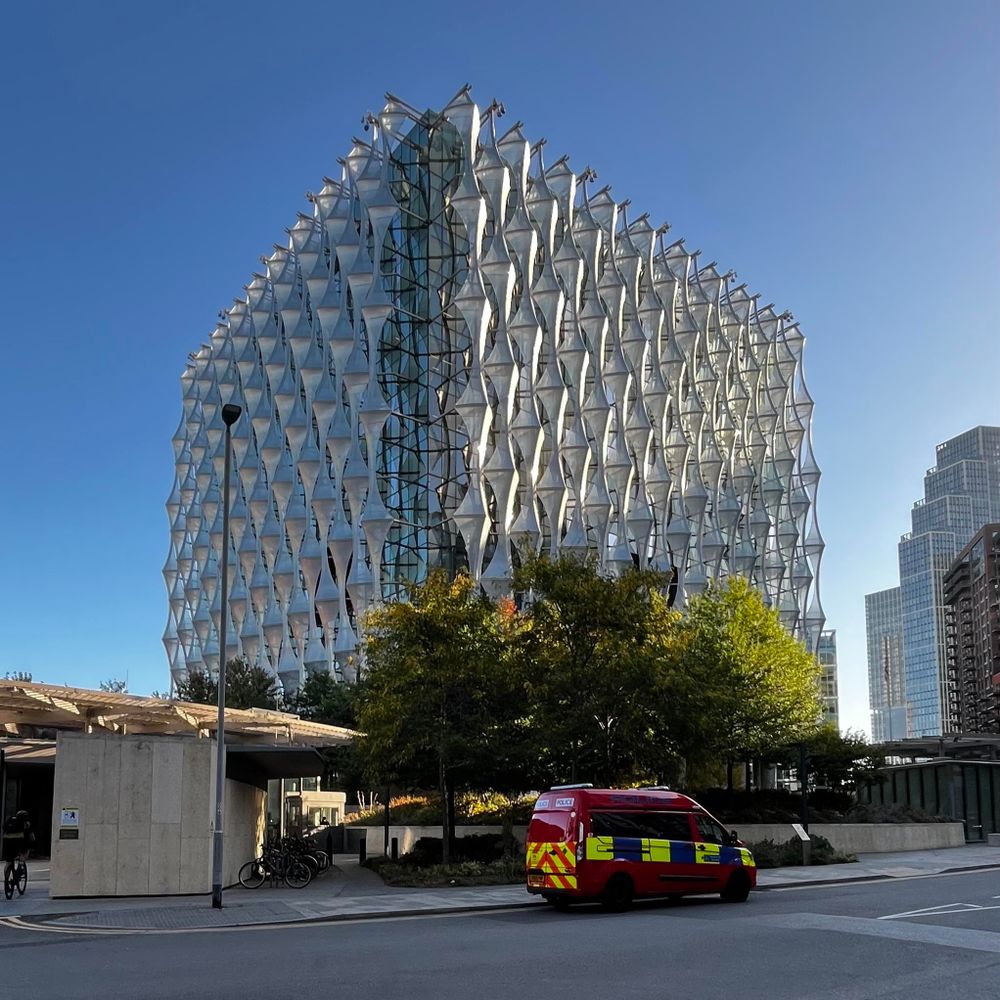 A modern, architecturally intricate building with a unique geometric facade is seen against a clear blue sky. In the foreground, a red emergency vehicle is parked near landscaped greenery and a small structure.