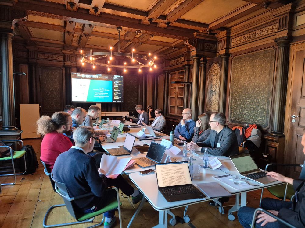 Menschen sitzen mit Laptops an einem großen Tisch in einem Raum. Vorne im Raum steht ein großer Monitor.