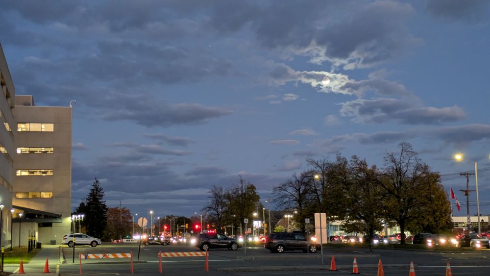 A parking lot with a large building on the left and a main road on the right. It the sky the moon is visible behind some light clouds