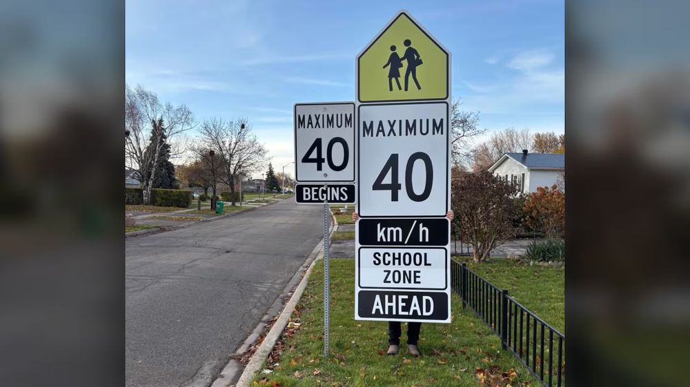 A normal sized "maximum 40 begins" sign on a pole beside a residential street, next to a 12-foot-high, maybe 3-foot-wide sign being held up my a man you cannot see because of the truly enormous sign he's holding, a veritable totem pole of road signage topped by a school zone image, and descending from that a huge wind-catching sail's worth of black and white sign, reading "Maximum 40 km/h - School zone - AHEAD". Notably, the sign blocks any view at all of the side of the road behind it, where, one assumes, children might be standing or walking, as there is no sidewalk.