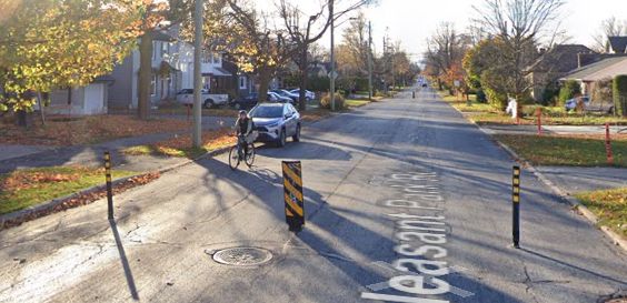 a google streetview of a street with temporary flex posts across it and a cyclist about to reach a point where they have to decide whether to go inside or outside the post that's in their way and might be too close to the curb to go outside