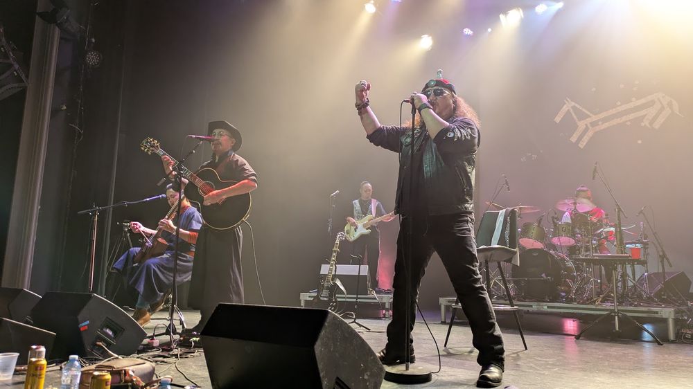 A band on stage. The three in front are a man in blue seated and playing a horesehead fiddle; a man in a long black coat and cowboy hat singing with a guitar; and a man in black jeans and a motorcycle vest with a Mongolian hat and aviator sunglasses, singing with his fist up and legs wide