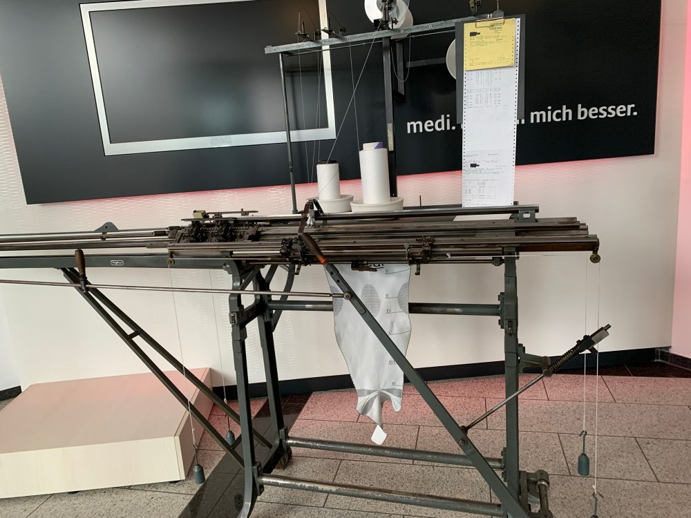 Eine alte Strickmaschine für Kompressionsstrumpfe, man sieht einen teilweise gestrickten Strumpf, Spulen mit weißem Garn, sowie am rechten Rand ein Massblatt 
Hinter der Strickmaschine eine dunkle Tafel mit der Aufschrift medi. (Schrift verdeckt) mich besser