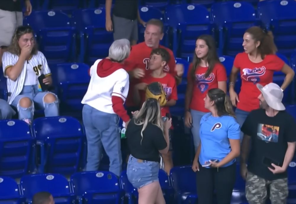 A gray-haired lady in baseball stands grabbing a father, who was hugging his son, & appears to berate the father while the son & other fans look on.