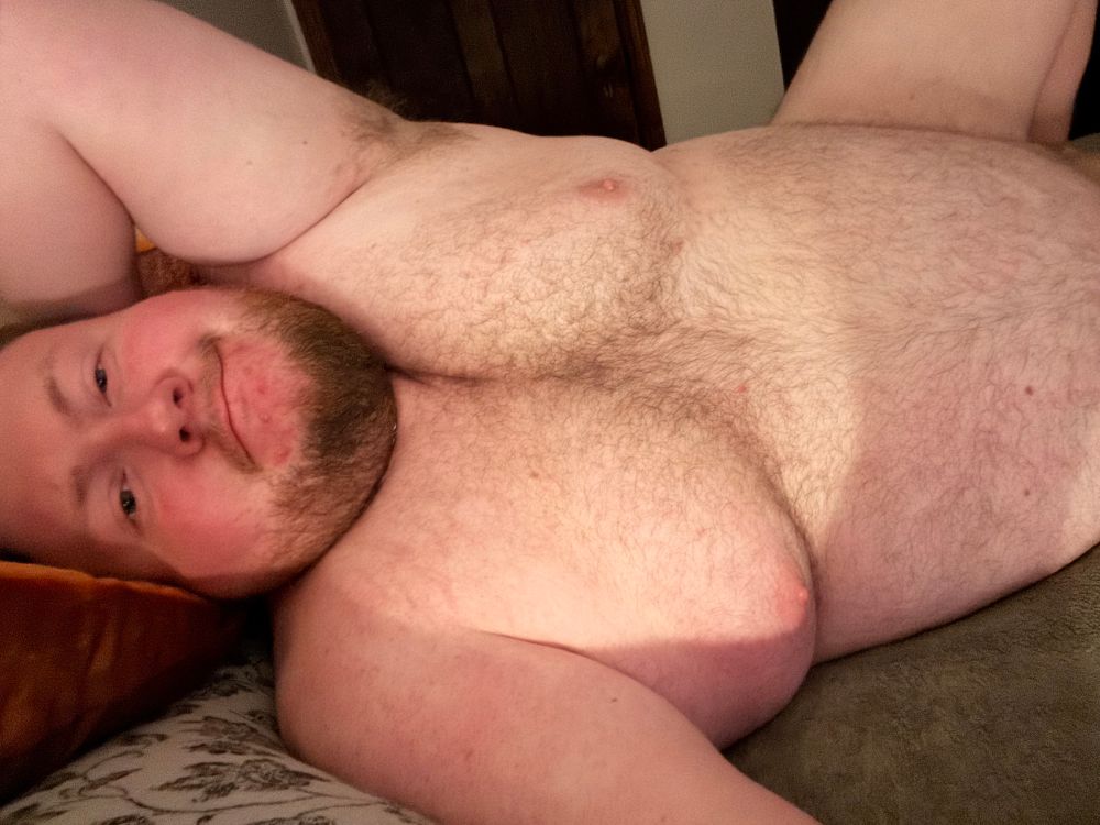 A chubby white bearded man laying on his side, one hand behind his head 