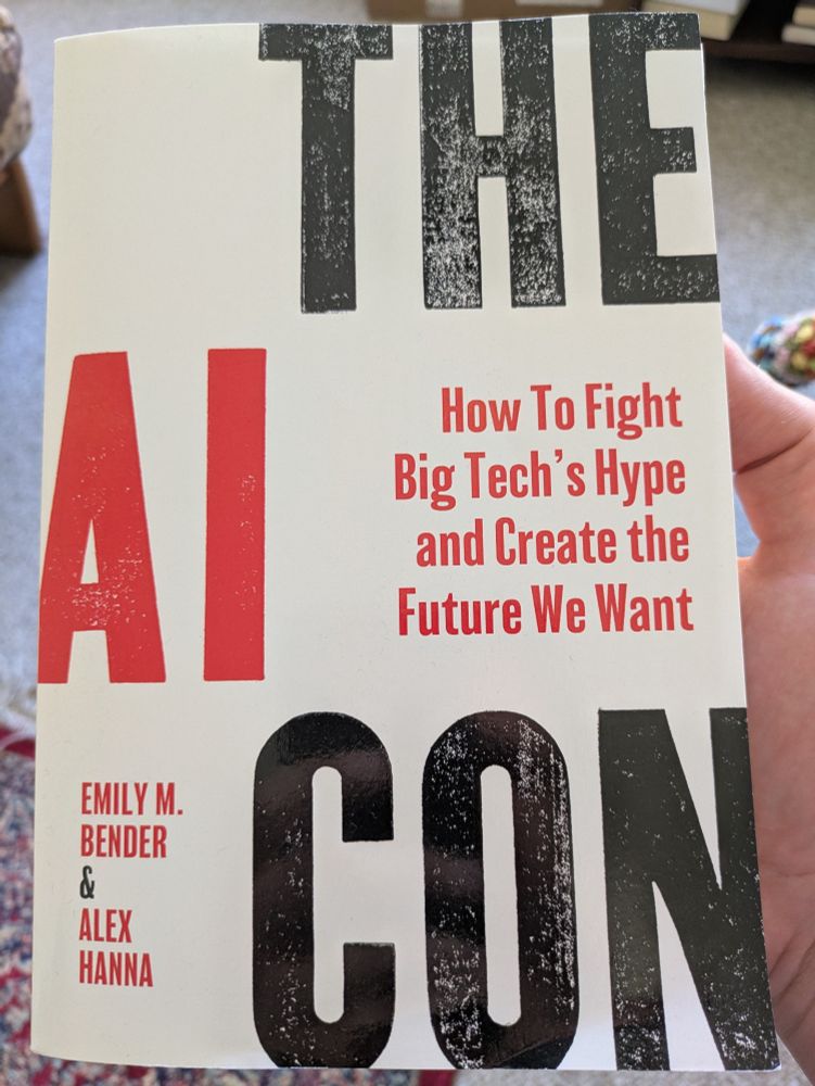 A hand holding a book titled "The AI Con: How to Fight Big Tech's Hype and Create the Future We Want," by Emily M. Bender and Alex Hanna