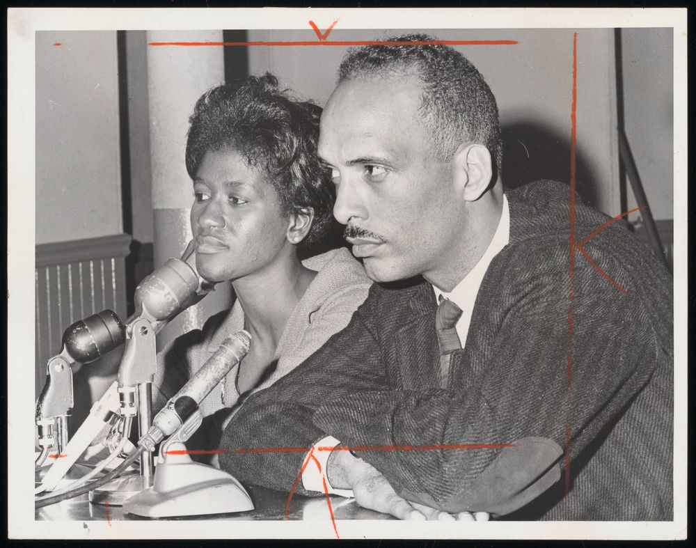 Ellen Jackson and Albert Raby seated at table with microphones addressing seeming to be listening to someone out of the frame. Raby is wearing a striped suit and his arms are crossed resting on the table while he raises an eyebrow. Ellen Jackson is resting her face on her hand with short curled hair while she listens. 