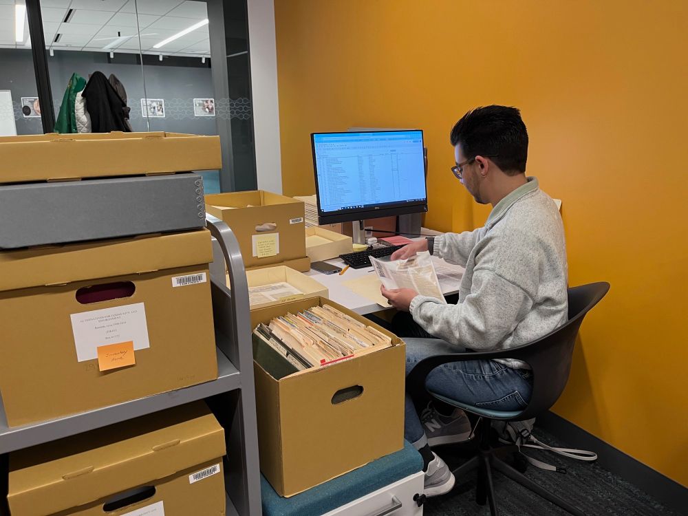 Person organizing papers into folders at a desk with a computer screen displaying a spreadsheet, surrounded by boxes full of files and a cart of boxes