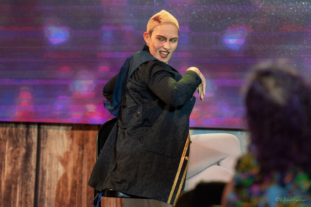 Polly Cocktail, in drag as Chauncey Badminton, sits on stage facing away from the audience and looking back over their shoulder.