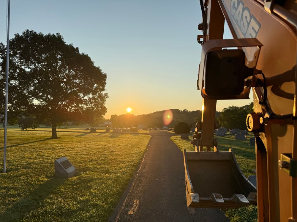 A serene sunrise over a cemetery, casting long shadows across the grass. A large tree stands to the left, and a CASE excavator with its bucket lowered is visible on the right, partially in frame. The sun is just above the horizon, bathing the scene in a golden glow along a paved path lined with gravestones. 