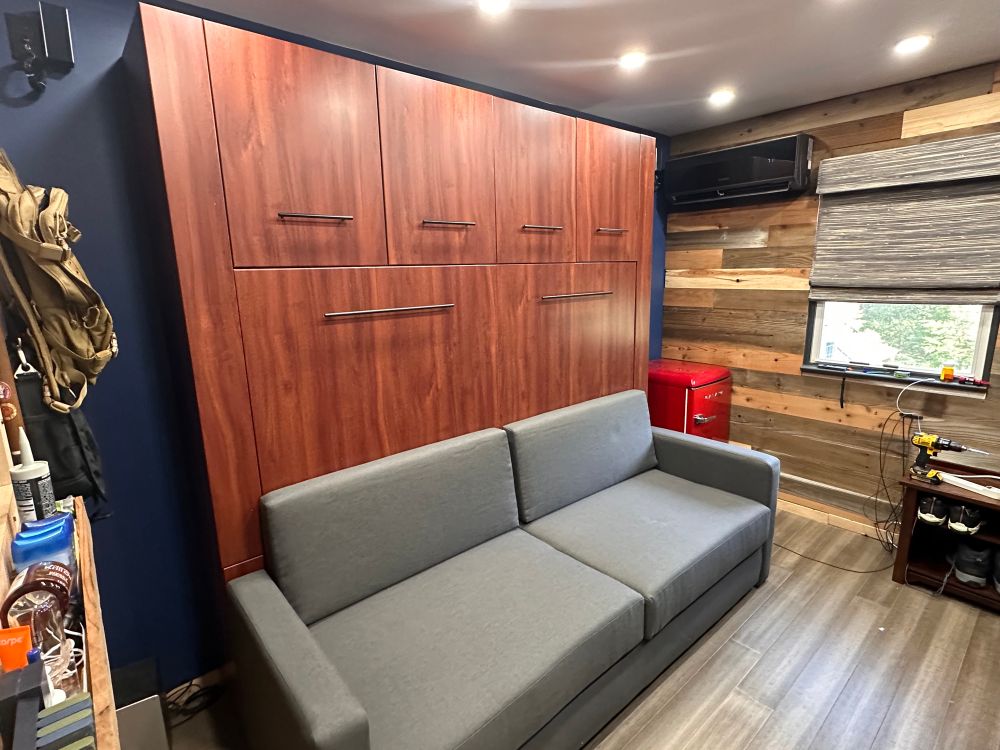A compact, modern room features a wall bed (Murphy bed) with a rich wood finish and black handles, folded up behind a gray sofa. The room has wood-paneled walls, recessed ceiling lights, a red mini fridge, a black mini-split A/C unit above the window, and organized tools and accessories on shelves and a small bench nearby.
