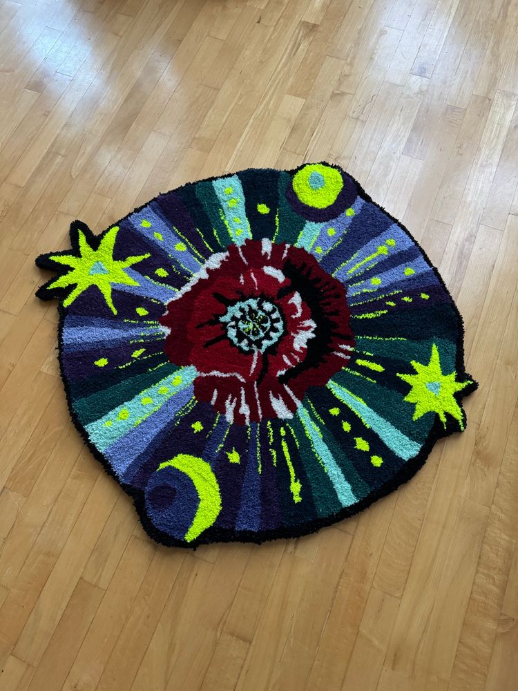 A rug depicting a poppy radiating out purples and greens into a celestial circle of stars and moons. It lays flat at an angle on a wood floor.