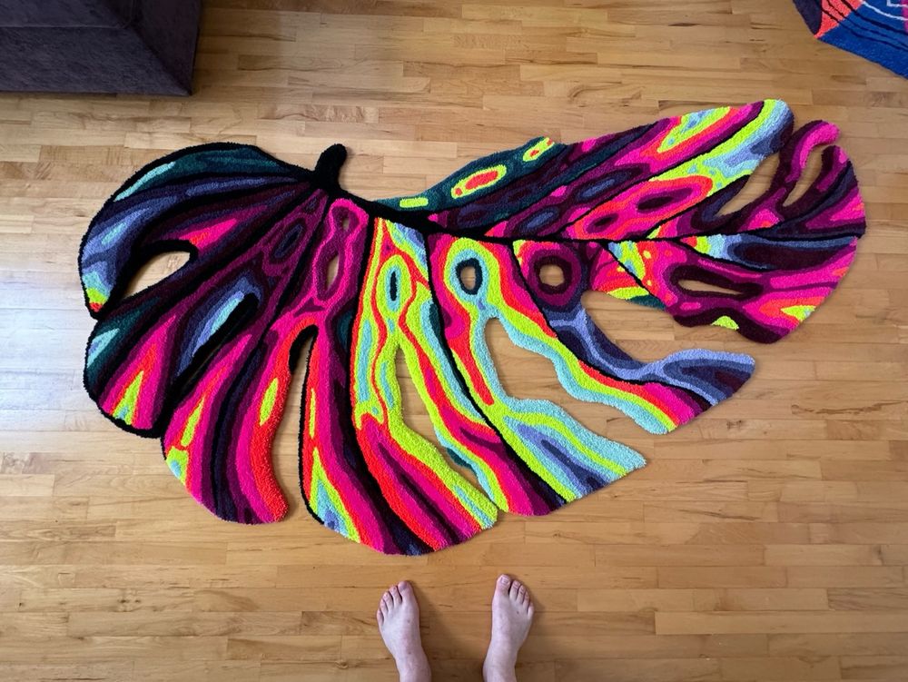 A monstera leaf rug made out of bold neon colors. Two feet are shown at the bottom to highlight the piece’s scale, about 6 feet across.