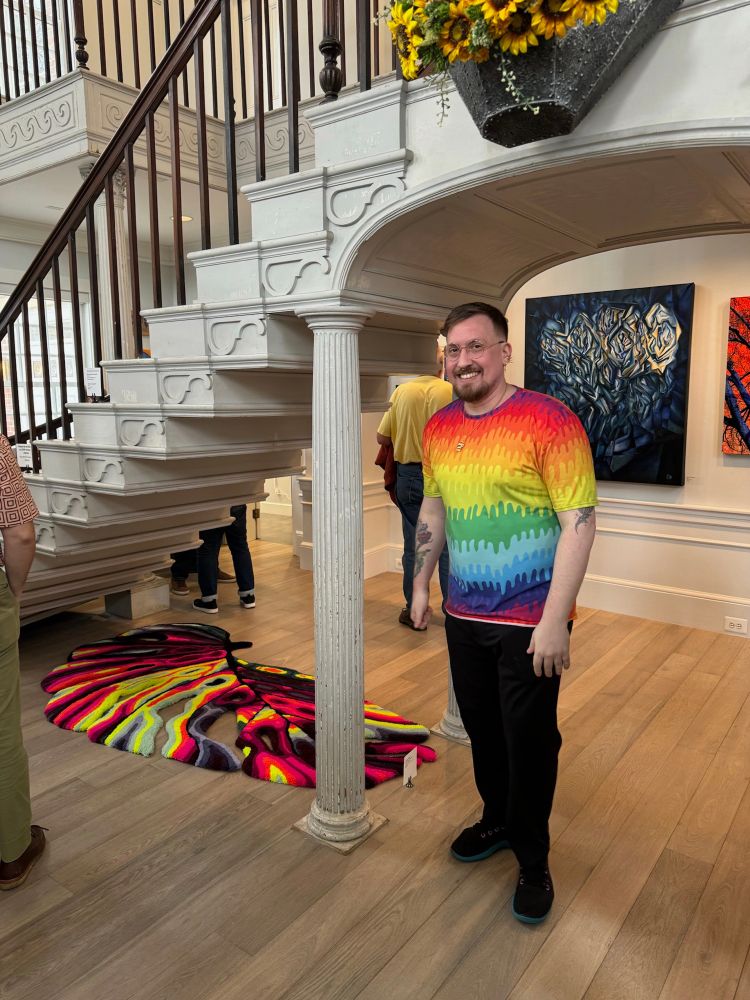 Quintin, the artist, stands in front of his work being featured at Somerville Museum. He’s wearing a rainbow drip tshirt and smiling. His piece is a monstera rug in his typical neon rainbow style and it sits under a staircase surrounded by paintings and other works.