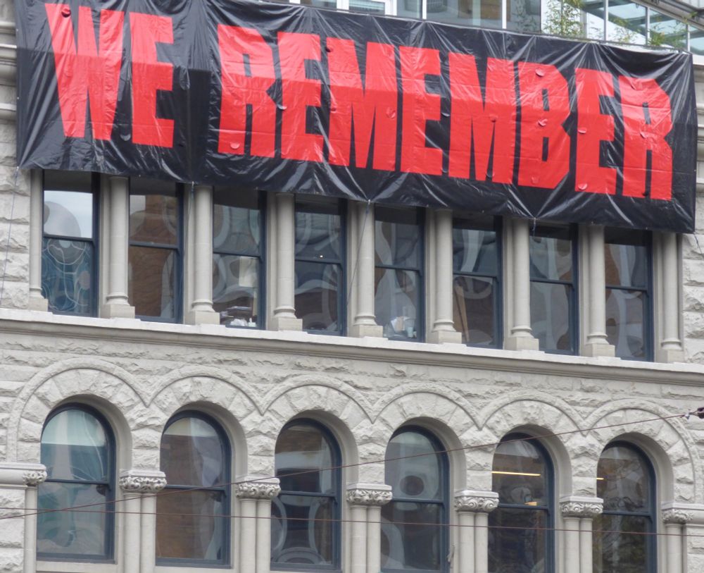 Photo of large Remembrance Day banner, displayed on the side of a building visible from Victory Square in Vancouver, B.C. "WE REMEMBER" is written in red capital letters on black. 