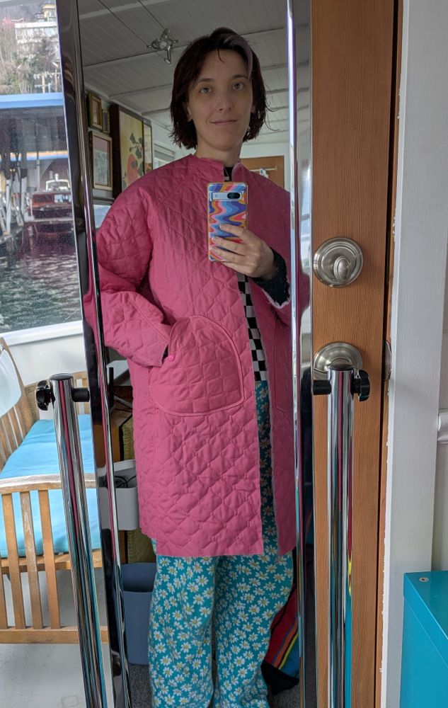 Al, a white woman with chin length dark hair,  is taking a mirror selfie. Shes wearing an unfinished pink quilted coat and blue floral pajama pants. She has her hand in the coat's heart-shaped pocket.