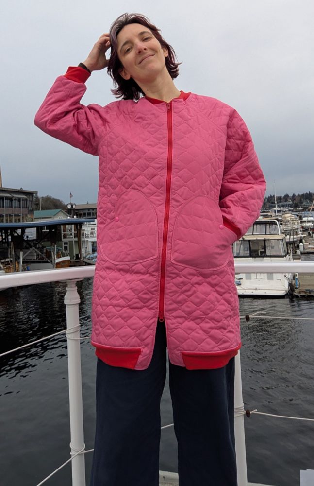 Al, a white woman with dark hair, smiles at the camera. She is wearing a pink quilted coat with red cuffs, collar, zipper and hem band. The coat has heart shaped patch pockets.