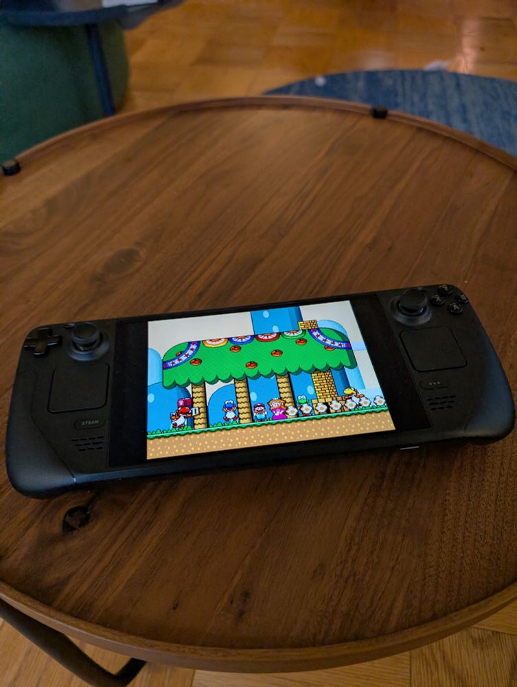 A steamdeck with the ending of super Mario world on a round walnut coffee table with black legs and a parque floor and a blue blue rug. 