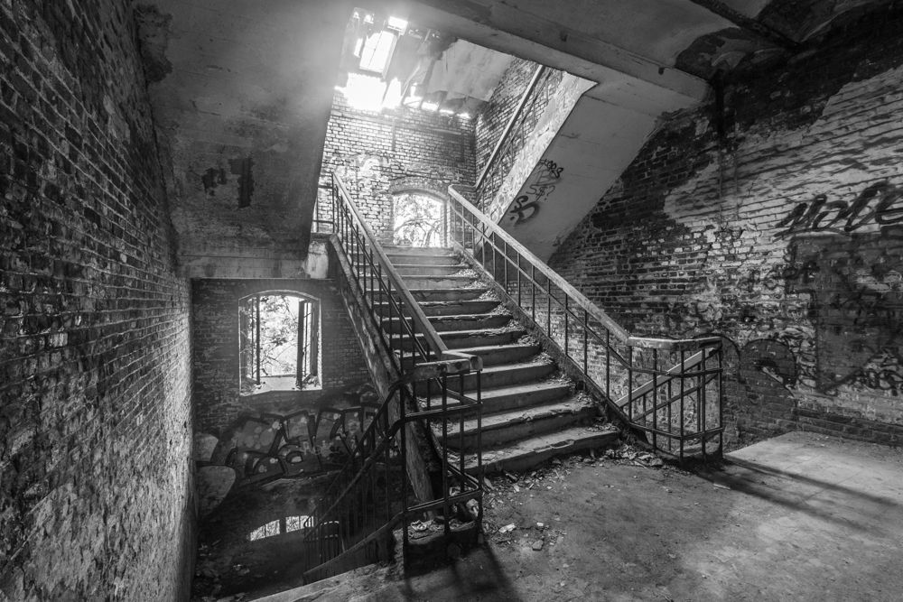 Black and white picture of a staircase in the abandoned fortress of chartreuse near Liège. Decay is visible, the roof is partially gone and the windows are broken. Left by the military more than 40 years ago, now a destination for urban explorers.