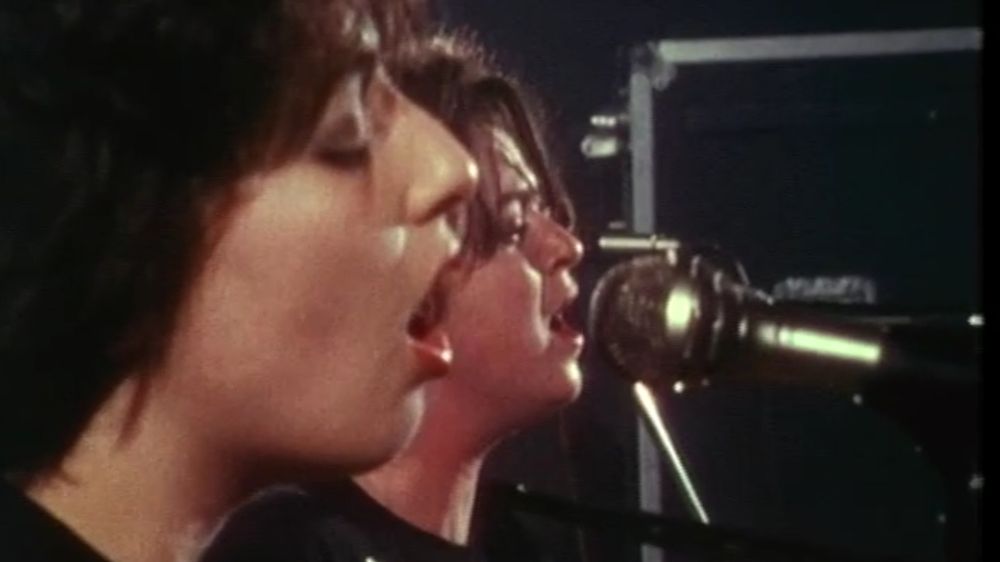 Zwei Sängerinnen sind zu sehen, aus naher Distanz seitlich aufgenommen. Ihre Münder sind geöffnet, sie singen in zwei Mikrofone, eins davon ist im Bild zu sehen. Es handelt sich um einen Screenshot aus einem Dokumentarfilm von 1989, Rachel Collins und Nicky Rushton waren die Band ‚And all because the lady loves‘ auf Tournee durch die Schweiz. Für, mit ihren Stimmen schlägt mein Herz.
