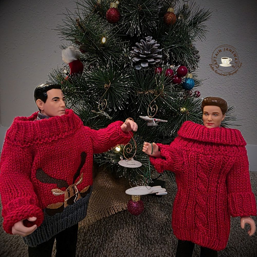 Action figures of Chakotay and Janeway both wearing holiday sweaters. There is a Christmas tree behind them. Janeway is hanging a Voyager ornament on the tree and Chakotay is hanging an Enterprise-D ornament. 