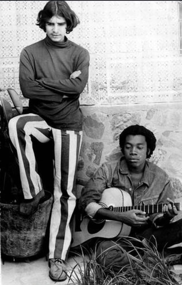 Lo Borges standing, wearing the coolest striped pants ever, with Milton Nascimento seated, holding a guitar 