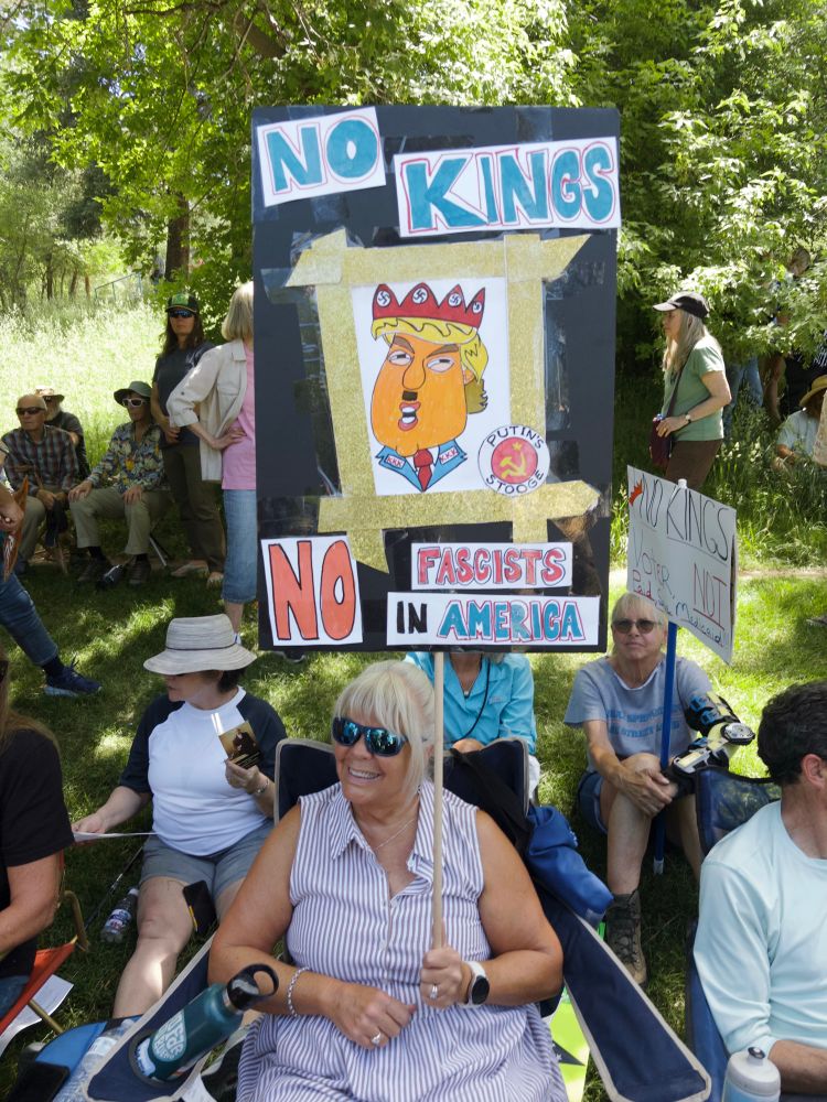 Woman with a sign: no kings, no fascists in America. With a picture of chubby orange guy with a crown and a Hitler mustache