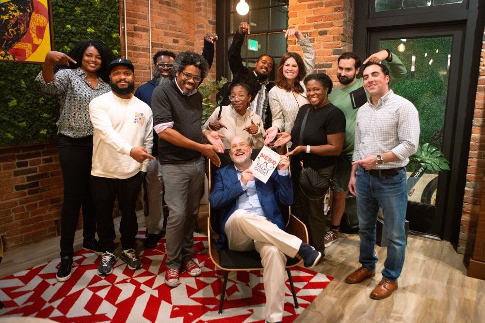 A group of smiling professionals standing around one person sitting in the middle on an easy chair. He's holding up a book and they're all pointing at his book.