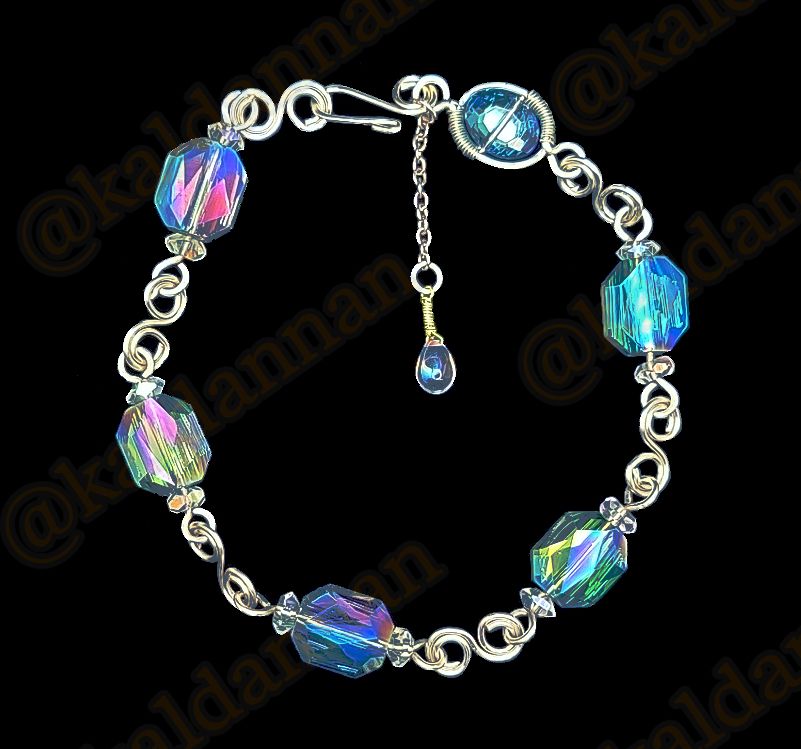 A silver wire bracelet atop a black background. There are six segments of blued, faceted glass on wire with loops on either end, connected with silver wire twisted into S shapes. A small briolette charm featuring a clear glass drop hangs from the base of the curved hook clasp by a short length of silver chain.
