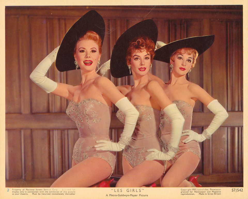 Poster for "Les Girls" (George Cukor, director, 1957) showing Mitzi Gaynor, Kay Kendall and Taina Elg in sparkly showgirl corsets, long white evening gloves, diamond bracelets and earrings and black picture hats. Each has one hand on her hip and the other on her hat.