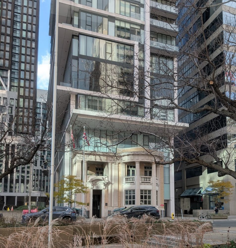 A small beige- and cream-coloured two-story facade with classical columns, loggias and porticos, installed at the base of a large, modern, glass and concrete building.