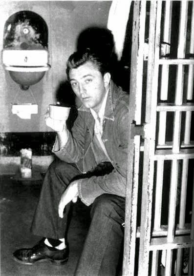 Photo of Robert Mitchum sitting on his bunk in his jail cell, holding a cup of coffee, 1948.