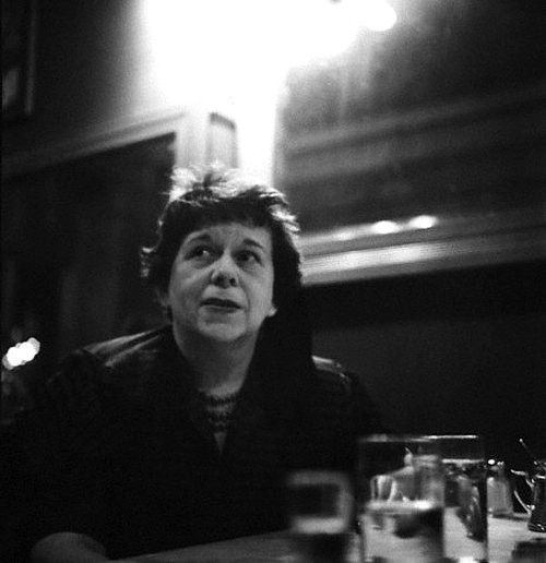 A middle-aged woman with short hair and dark lipstick, seated at a table in a restaurant, looking up with a quizzical expression. Black and white photo of Dawn Powell at the Lafayette Hotel, New York, 1950s, by Genevieve Naylor. 