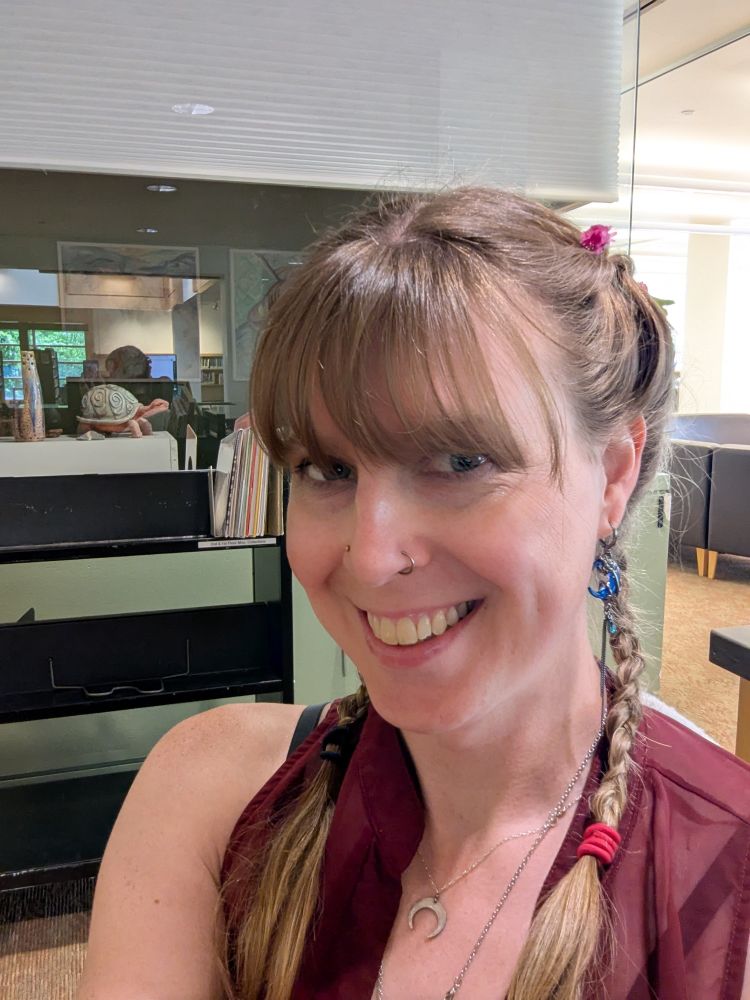 Picture of a smiling woman in a red top with a moon necklace and earrings. Her hair is in two braids and flowers are woven into the top of the braid.