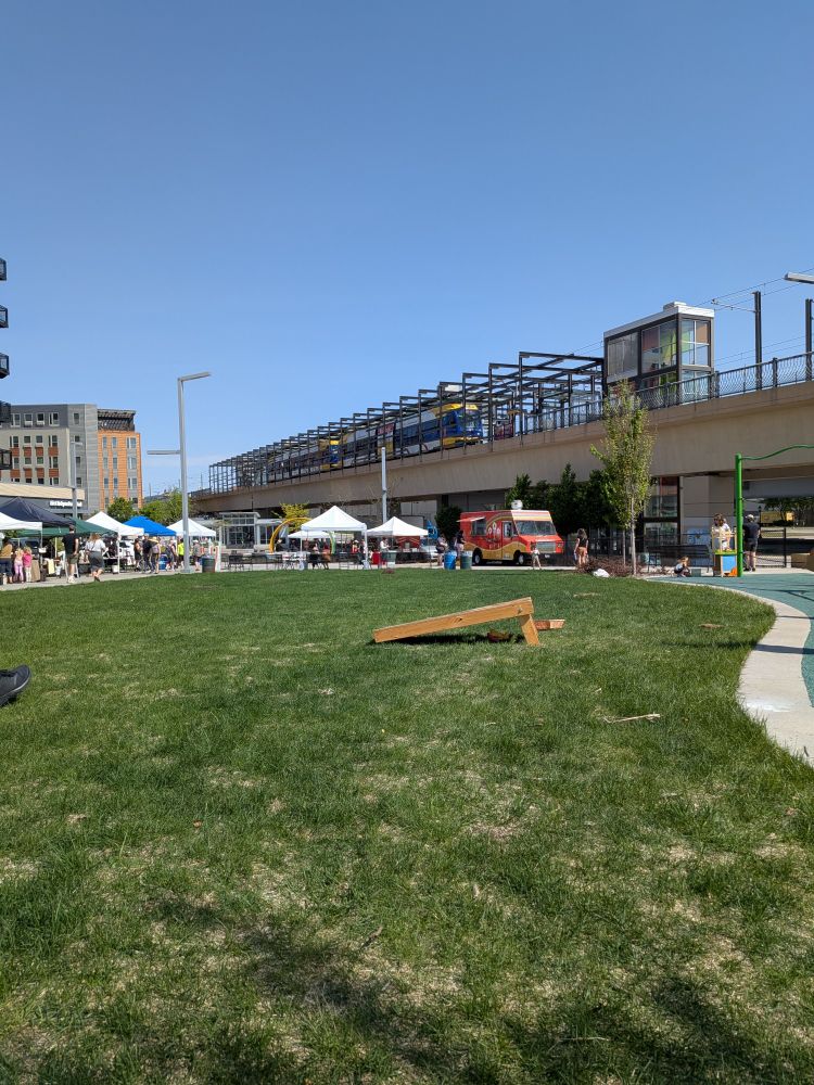 A picture of the Midtown Farmer's Market in Minneapolis. There is green grass, a bean bag toss board, a food truck, several vendor tents and an elevated light rail track and train. 