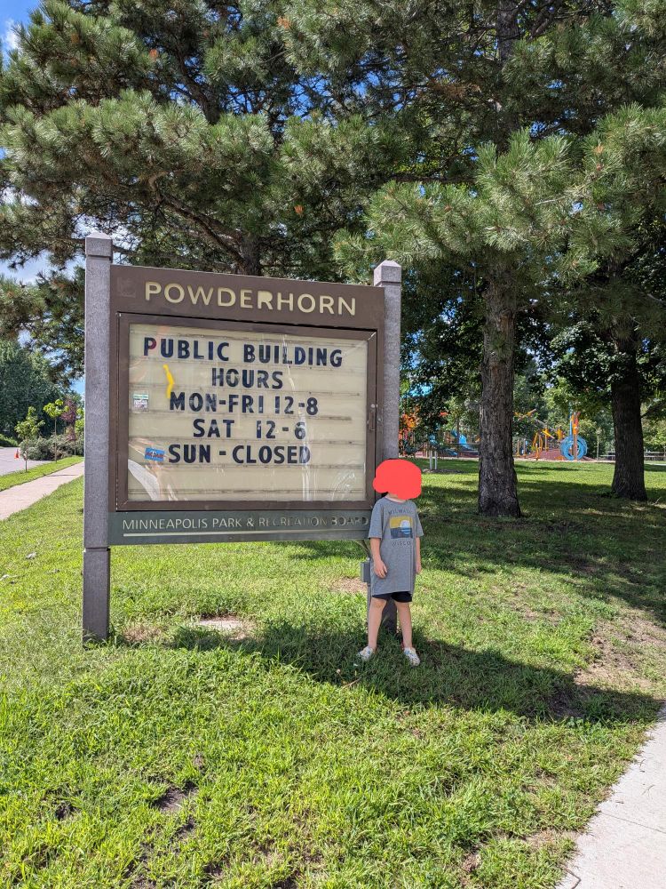 A kid in front of the Powderhorn Park sign, lots of green trees and grass all around.