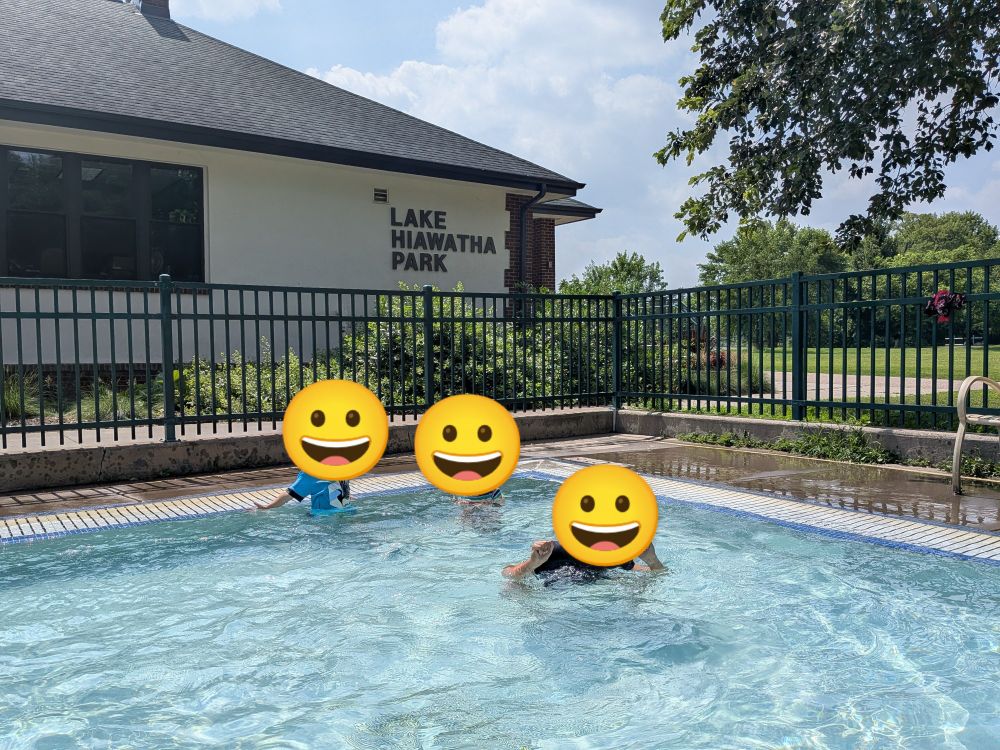 3 kids with their faces blocked out with smiley emojis. They're in a splash pad with a gate and park pavilion that says, 