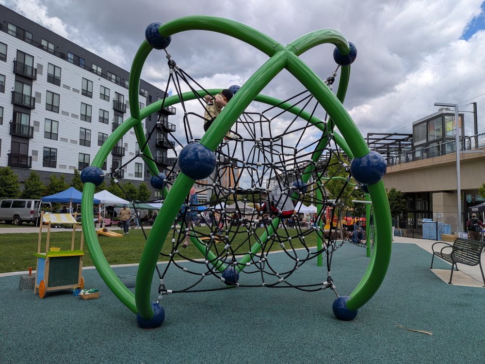 A green outdoor climbing play structure with a few kids on it. Behind that you can see vendor canopies, grass and benches. There's an apartment building and a raised light rail platform in the background. 