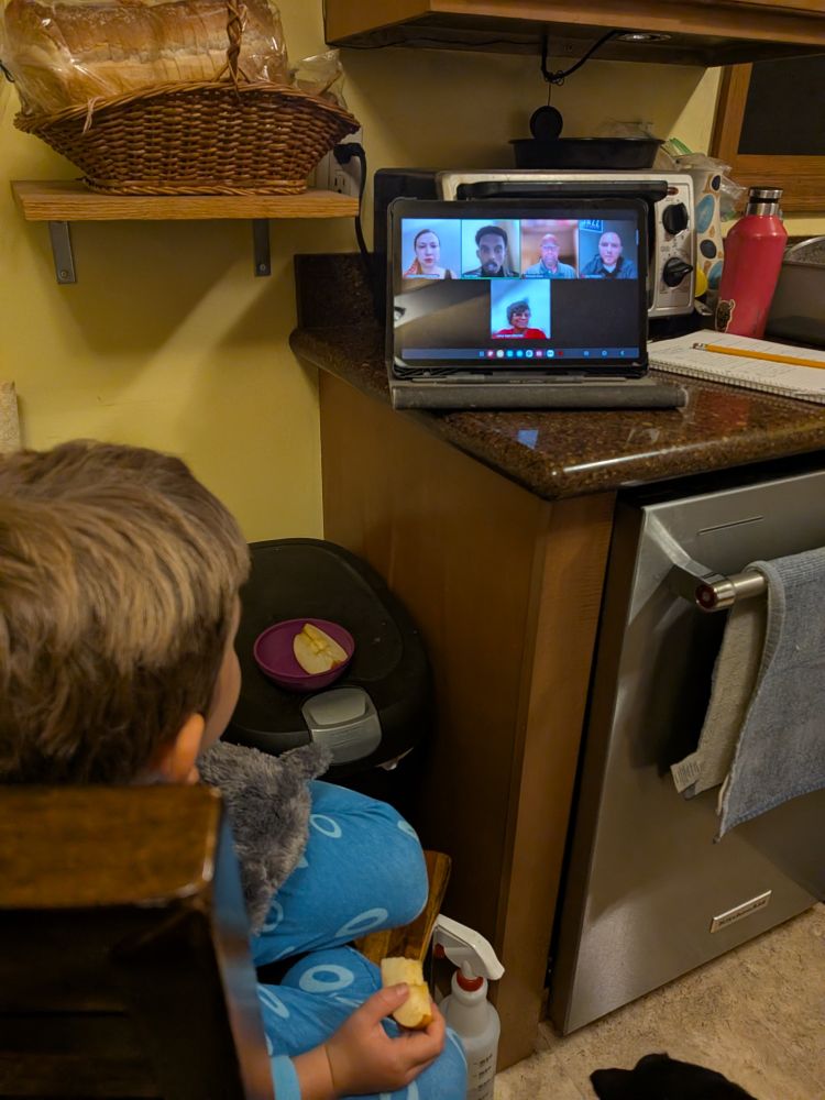 A tablet showing a Zoom call with Mayoral candidates Omar Fateh, Dewayne Davis, Jazz Hampton and MFE representatives Catina Taylor and Natasha Docktor. The tablet is on a kitchen counter, a young kid in pajamas is watching the screen with an apple slice in his hand. 