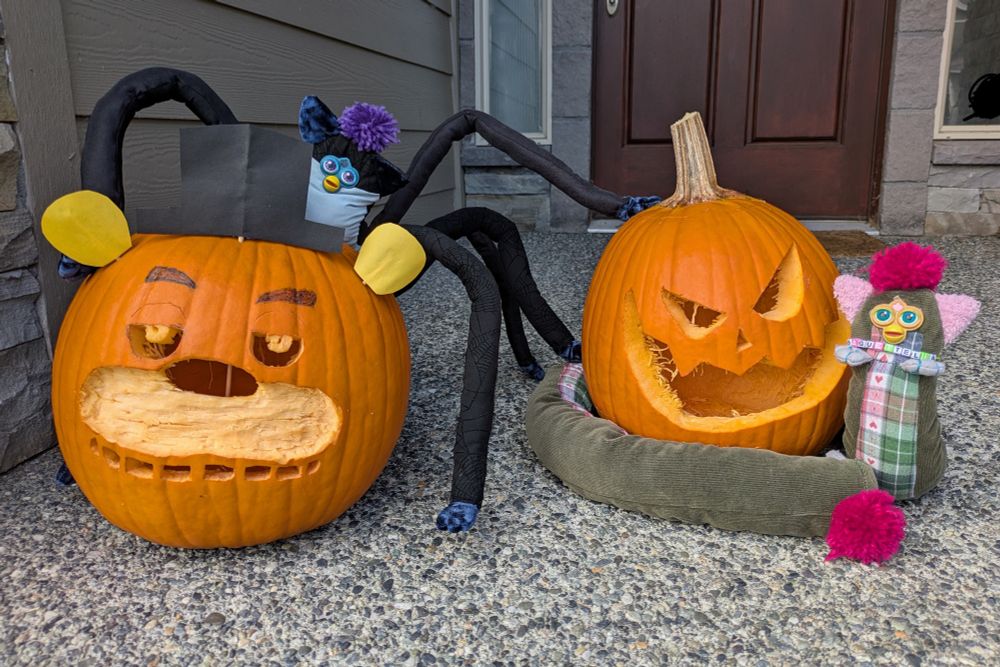 Two customized furbys sit on a porch showing off carved pumpkins. On the right, Baguettella, a green and pink long furby, is coiled around a classic jack-o-lanturn, while on the left Yux, a black and blue spider with a furby's face and furby paws, is lurching over a pumpkin carved with the face of Freddy Fazbear from Five Nights at Freddy's, complete with a paper top hat and ears.