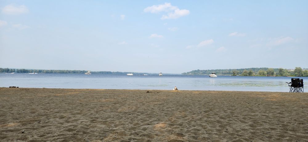 A picture of Petrie Island beach showing the wide sandy beach, the open water with some boats on it, and forested hills and riverbanks in the distance.